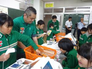 セブンイレブンの制服を着た小学生がレジの体験をしています。