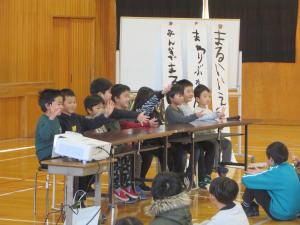 1年生が椅子に座り、2学期の思い出を習字で書いたものを全校に見せながら発表している様子。