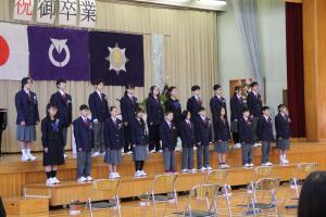卒業生21名がステージ上に並んび、在校生や保護者にむけて呼びかけと歌を歌う様子。